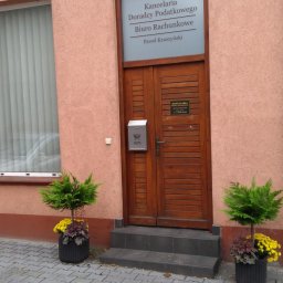 KANCELARIA DORADCY PODATKOWEGO BIURO RACHUNKOWE Paweł Kruszyński - Wejście do kancelarii z drewnianymi drzwiami, skrzynką pocztową i roślinami w donicach, tabliczka informacyjna nad drzwiami z napisem 'Kancelaria Doradcy Podatkowego Biuro Rachunkowe Paweł...