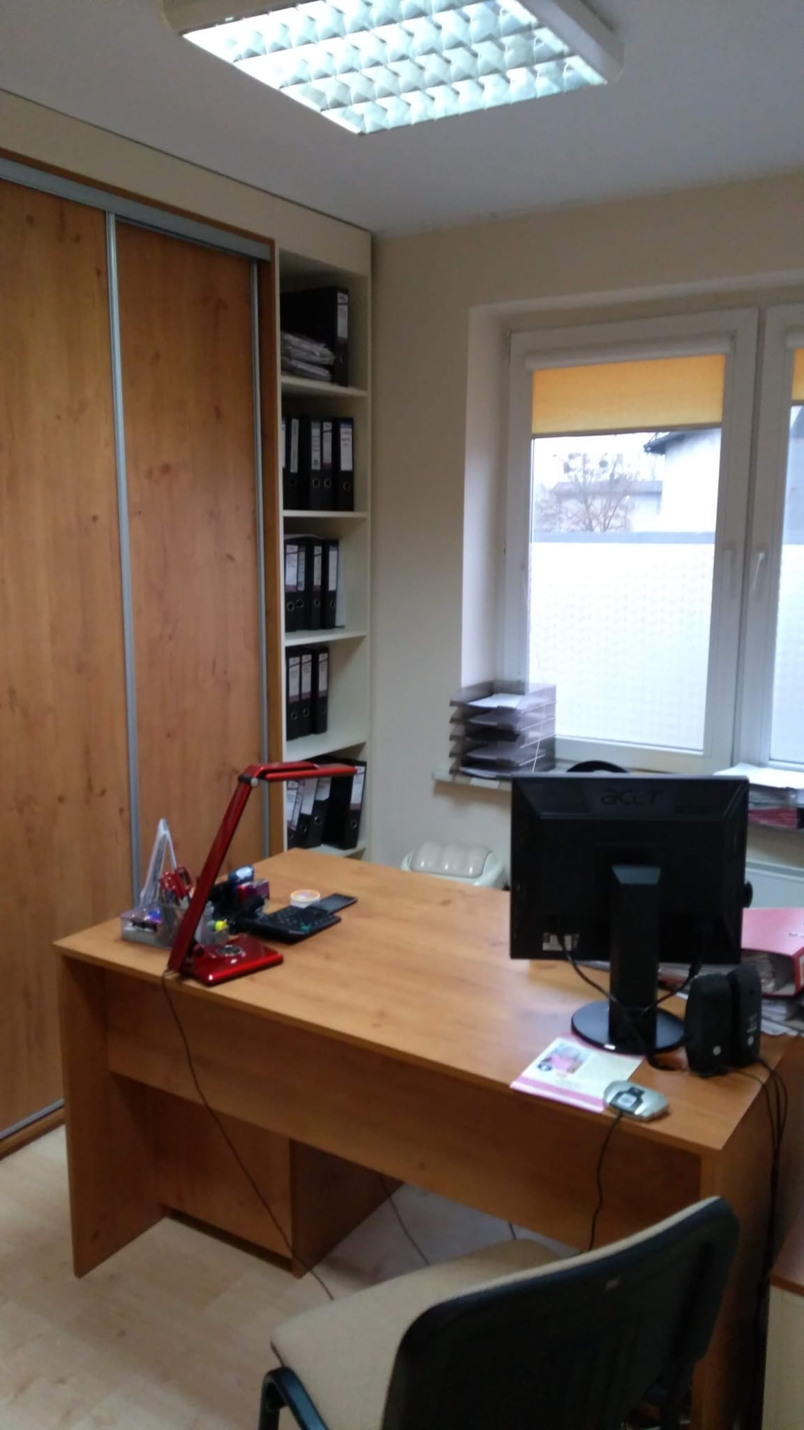 Biuro w Brodnicy z biurkiem, komputerem, lampką i regałem pełnym segregatorów. Widok na uporządkowane miejsce pracy.
