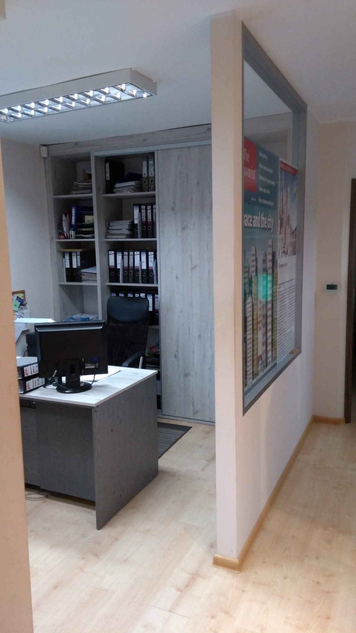 Biuro z biurkiem, komputerem i regałem wypełnionym segregatorami, widoczne plakaty na ścianie.