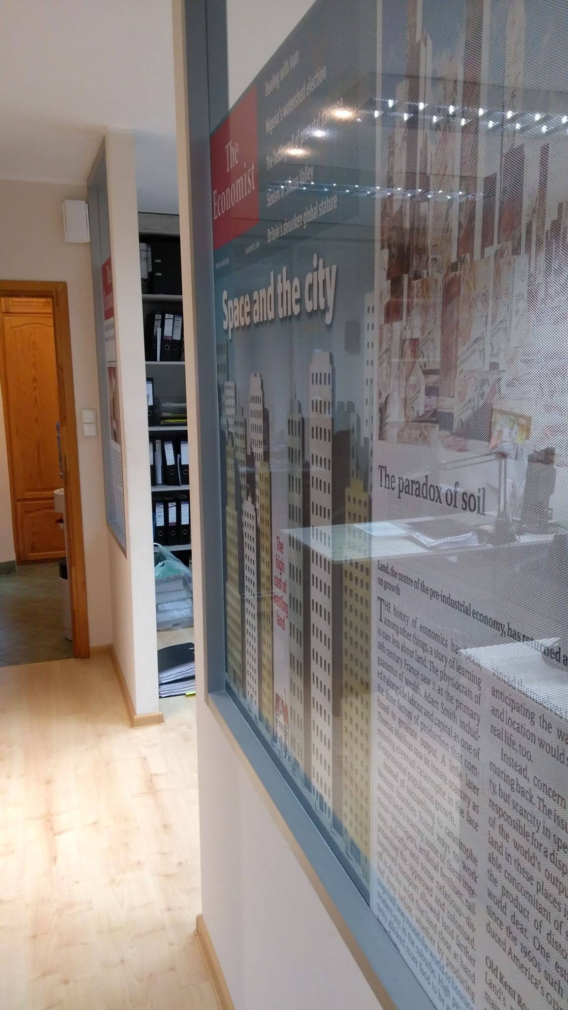 Biuro z regałami na dokumenty; na pierwszym planie ściana z naklejonymi artykułami z gazety The Economist o tematyce ekonomicznej i miejskiej.
