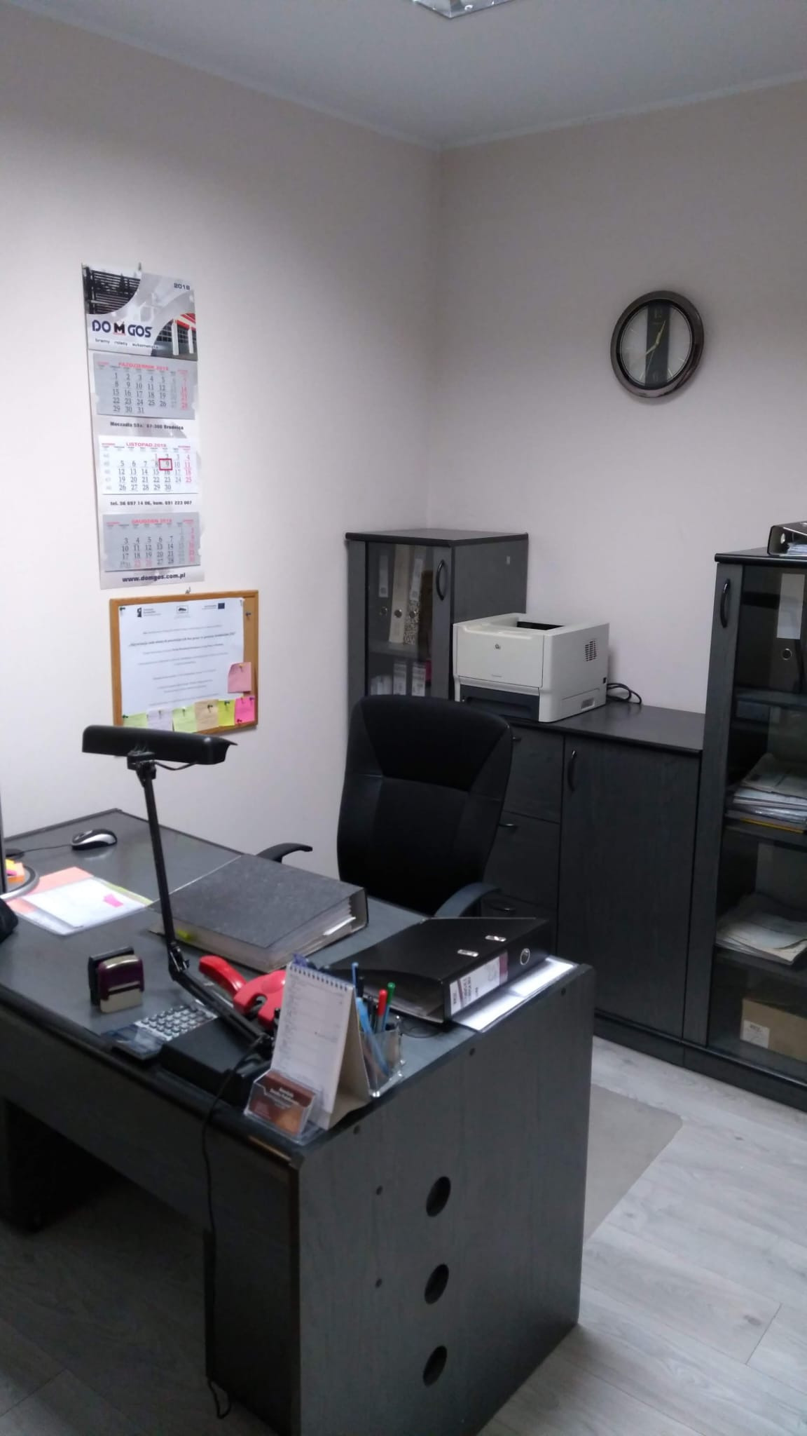 Biuro z czarnymi meblami, kalendarzem na ścianie i biurkiem z dokumentami, kalkulatorem i lampką.