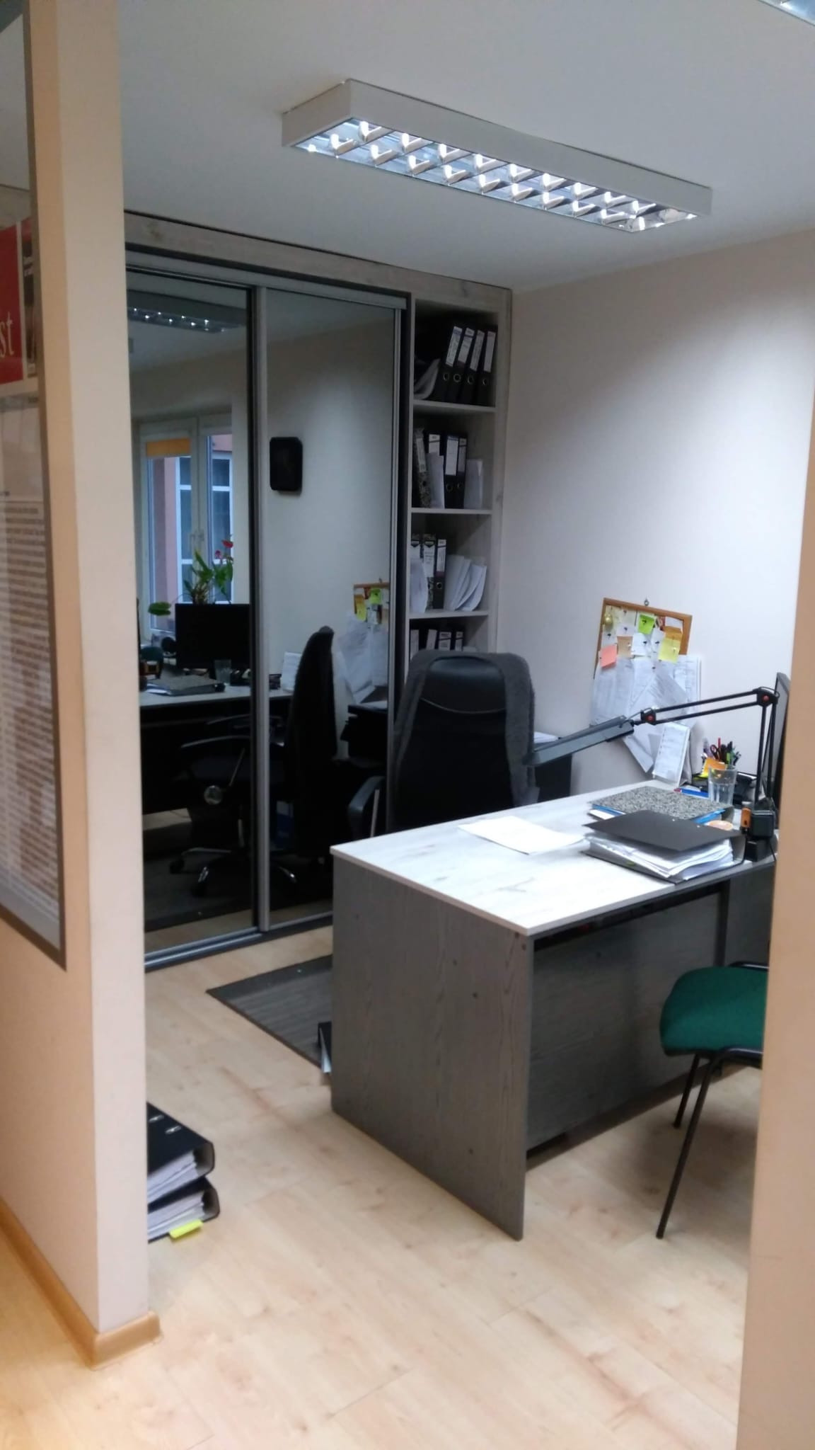 Wnętrze biura z biurkiem, krzesłem i regałem na dokumenty. Na biurku lampka, stos dokumentów i przybory biurowe. W tle widoczne lustrzane drzwi szafy i kolejne stanowisko pracy.