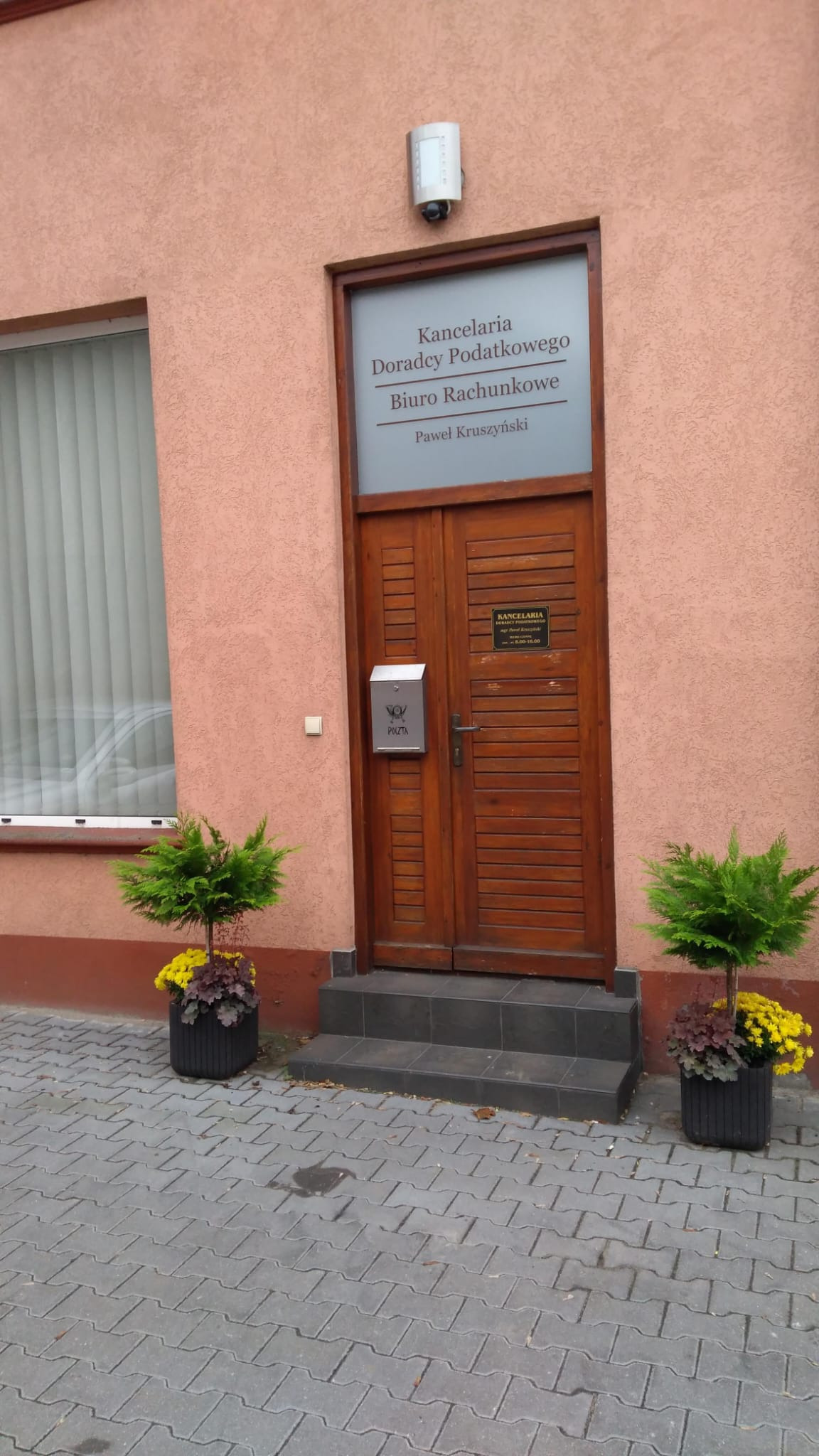 Wejście do kancelarii z drewnianymi drzwiami, skrzynką pocztową i roślinami w donicach, tabliczka informacyjna nad drzwiami z napisem 'Kancelaria Doradcy Podatkowego Biuro Rachunkowe Paweł...