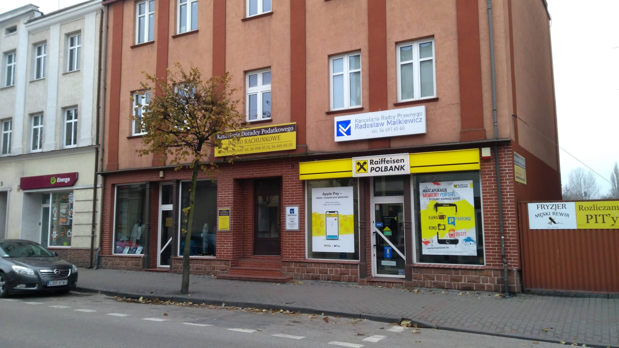 Budynek z biurami usługowymi, w tym Kancelaria Doradcy Podatkowego i Biuro Rachunkowe z żółtym szyldem, obok Raiffeisen Polbank i fryzjer męski, na ulicy z drzewem i zaparkowanym samochodem.