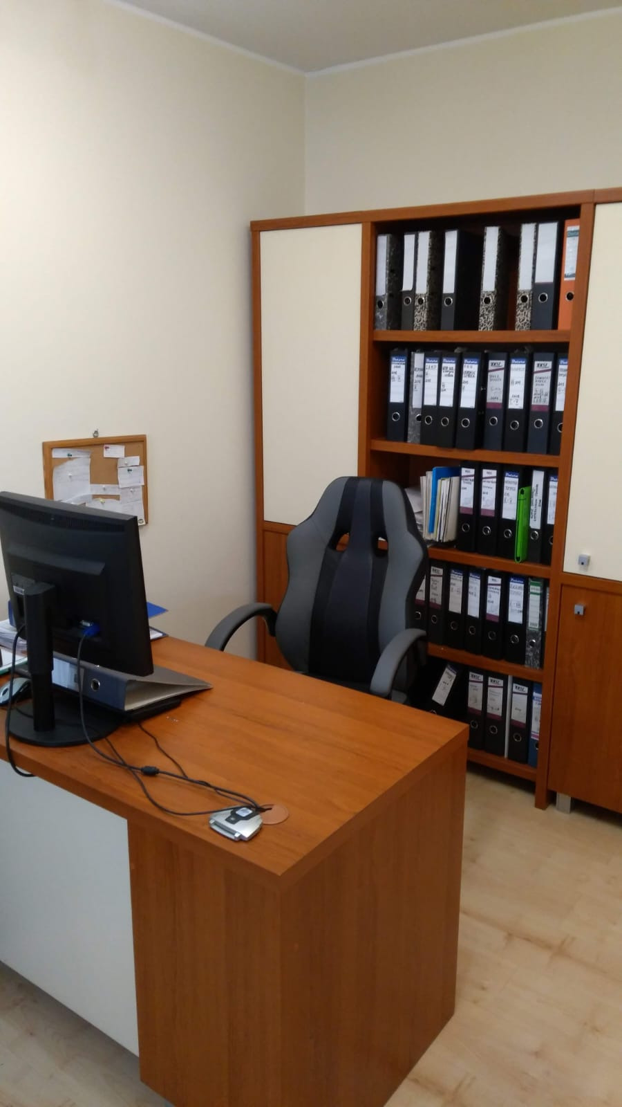 Biuro z biurkiem z drewnianym blatem, krzesłem biurowym w stylu gamingowym i regałem pełnym segregatorów z dokumentami.