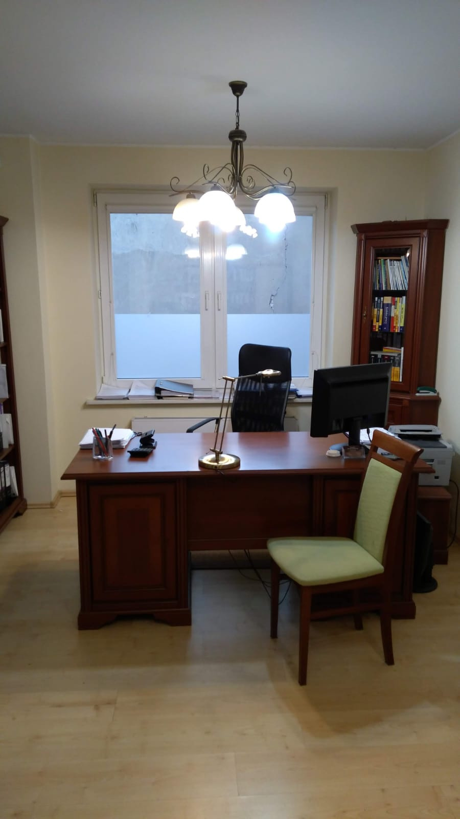 Biuro z drewnianym biurkiem, krzesłem z zielonym obiciem i regałem na książki, oświetlone lampą wiszącą z białymi kloszami.