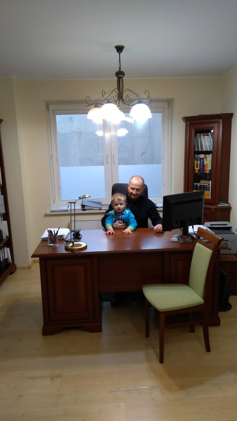 Mężczyzna siedzi przy biurku w biurze, trzymając małe dziecko na kolanach; w tle regał z książkami, komputer i lampa na biurku.