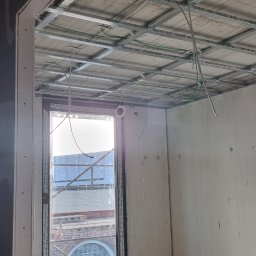 sucha_zabudowa.slowinski - Montaż metalowej konstrukcji sufitu podwieszanego w Chocianowicach, widoczne profile, przewody elektryczne i okno z widokiem na budynek z cegły.