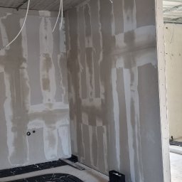 sucha_zabudowa.slowinski - Wnętrze w trakcie prac wykończeniowych: ściany pokryte wstępną warstwą gładzi na płytach gipsowo-kartonowych, widoczne instalacje elektryczne i rury ogrzewania podłogowego na surowym betonie.