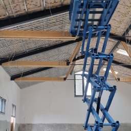 sucha_zabudowa.slowinski - Wnętrze w trakcie prac wykończeniowych: widok na ocieplony dach z drewnianą konstrukcją, niebieski podnośnik nożycowy w centrum kadru, surowe ściany z widocznymi śladami szpachlowania, okna i kable...