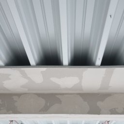 sucha_zabudowa.slowinski - Sufit z blachy trapezowej, widoczne mocowania, poniżej sufit z niedokończonymi gładziami, wystające przewody elektryczne i okno z widokiem na drzewa.