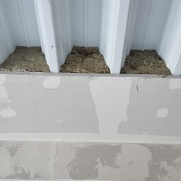 sucha_zabudowa.slowinski - Wnętrze w trakcie prac wykończeniowych: izolacja wełną mineralną pod metalowym dachem, płyty gipsowo-kartonowe na ścianie, widoczne przewody elektryczne w metalowych profilach.