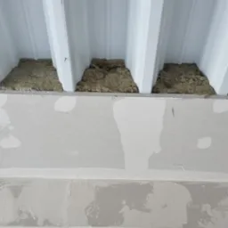 Wnętrze w trakcie prac wykończeniowych: izolacja wełną mineralną pod metalowym dachem, płyty gipsowo-kartonowe na ścianie, widoczne przewody elektryczne w metalowych profilach.