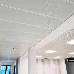 sucha_zabudowa.slowinski - Nowoczesny sufit podwieszany z paneli perforowanych w jasnym wnętrzu biurowym. Widoczne elementy oświetleniowe i fragmenty ścian w trakcie wykończenia.