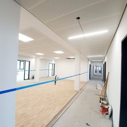 sucha_zabudowa.slowinski - Wnętrze w trakcie wykończenia: drewniana podłoga częściowo ułożona, sufit podwieszany z oświetleniem. W tle widoczne drzwi i narzędzia, niebieska taśma wyznacza obszar robót.