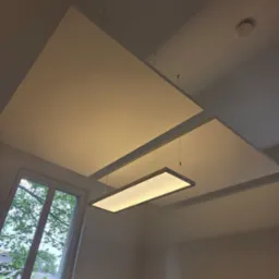 Nowoczesny sufit podwieszany z płyt w kształcie kwadratów, podświetlony wiszącą lampą. Widoczne okno z zielenią za szybą i czujnik dymu na suficie.