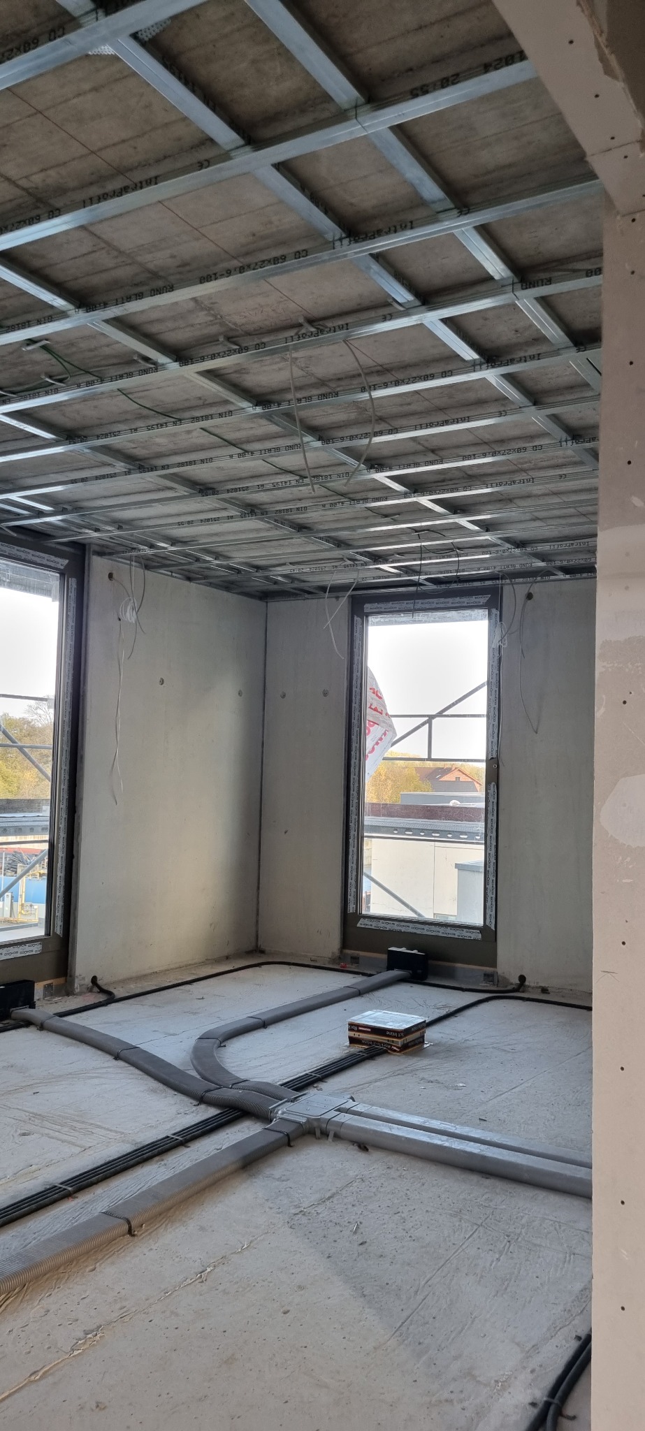 Wnętrze w trakcie prac wykończeniowych: widoczna metalowa konstrukcja sufitu podwieszanego z płytami, instalacja elektryczna w peszlach ułożona na surowej posadzce, ściany przygotowane pod gładzie,...