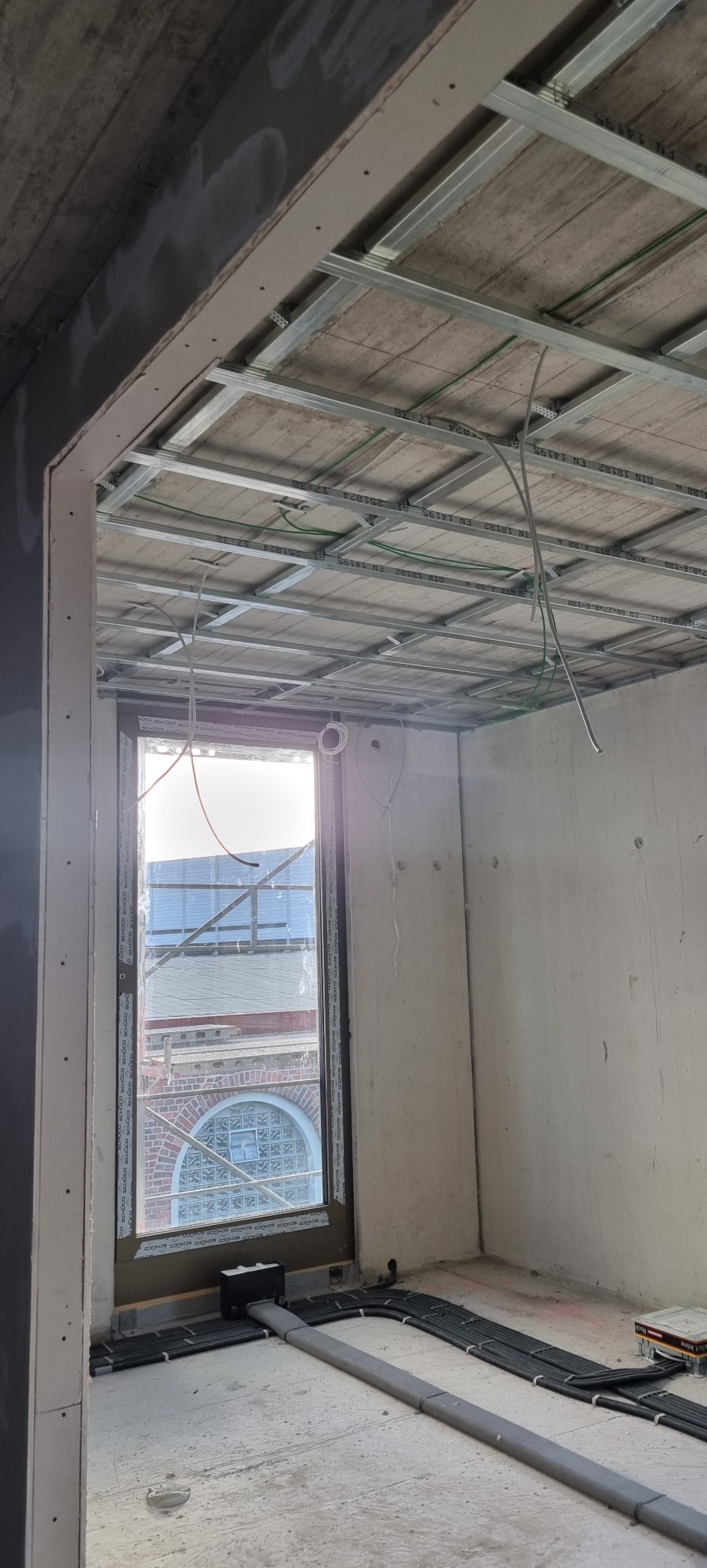 Montaż metalowej konstrukcji sufitu podwieszanego w Chocianowicach, widoczne profile, przewody elektryczne i okno z widokiem na budynek z cegły.