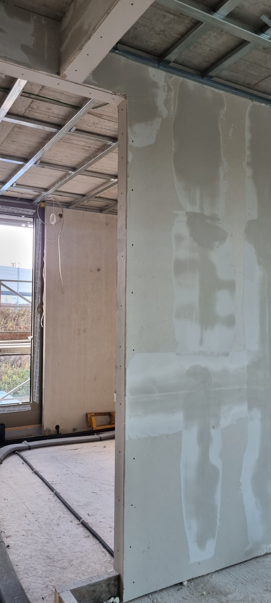 Wnętrze w trakcie prac wykończeniowych: widoczna konstrukcja sufitu podwieszanego, ściana z płyt gipsowo-kartonowych po szpachlowaniu, widok przez otwór drzwiowy na surowy pokój z oknem...