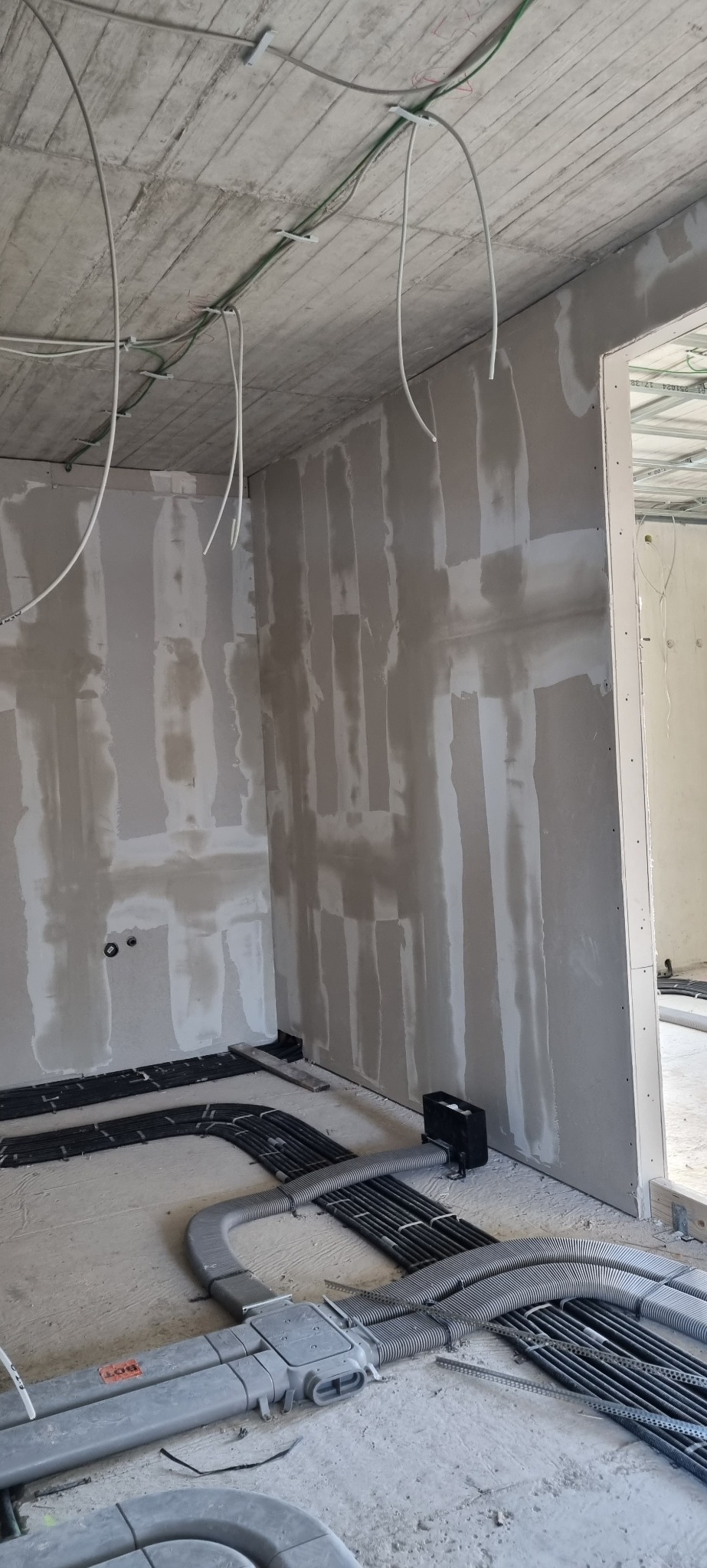 Wnętrze w trakcie prac wykończeniowych: ściany pokryte wstępną warstwą gładzi na płytach gipsowo-kartonowych, widoczne instalacje elektryczne i rury ogrzewania podłogowego na surowym betonie.