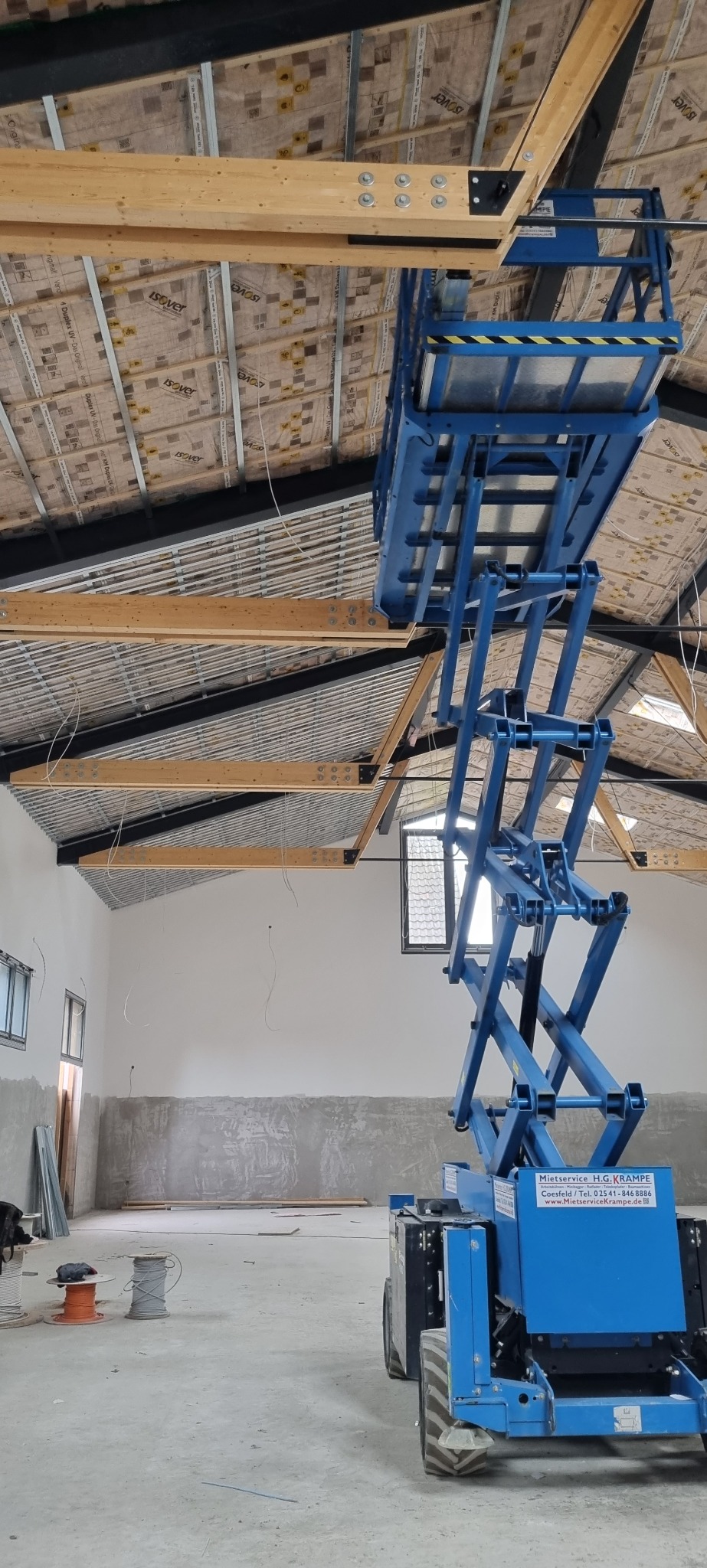 Wnętrze w trakcie prac wykończeniowych: widok na ocieplony dach z drewnianą konstrukcją, niebieski podnośnik nożycowy w centrum kadru, surowe ściany z widocznymi śladami szpachlowania, okna i kable...