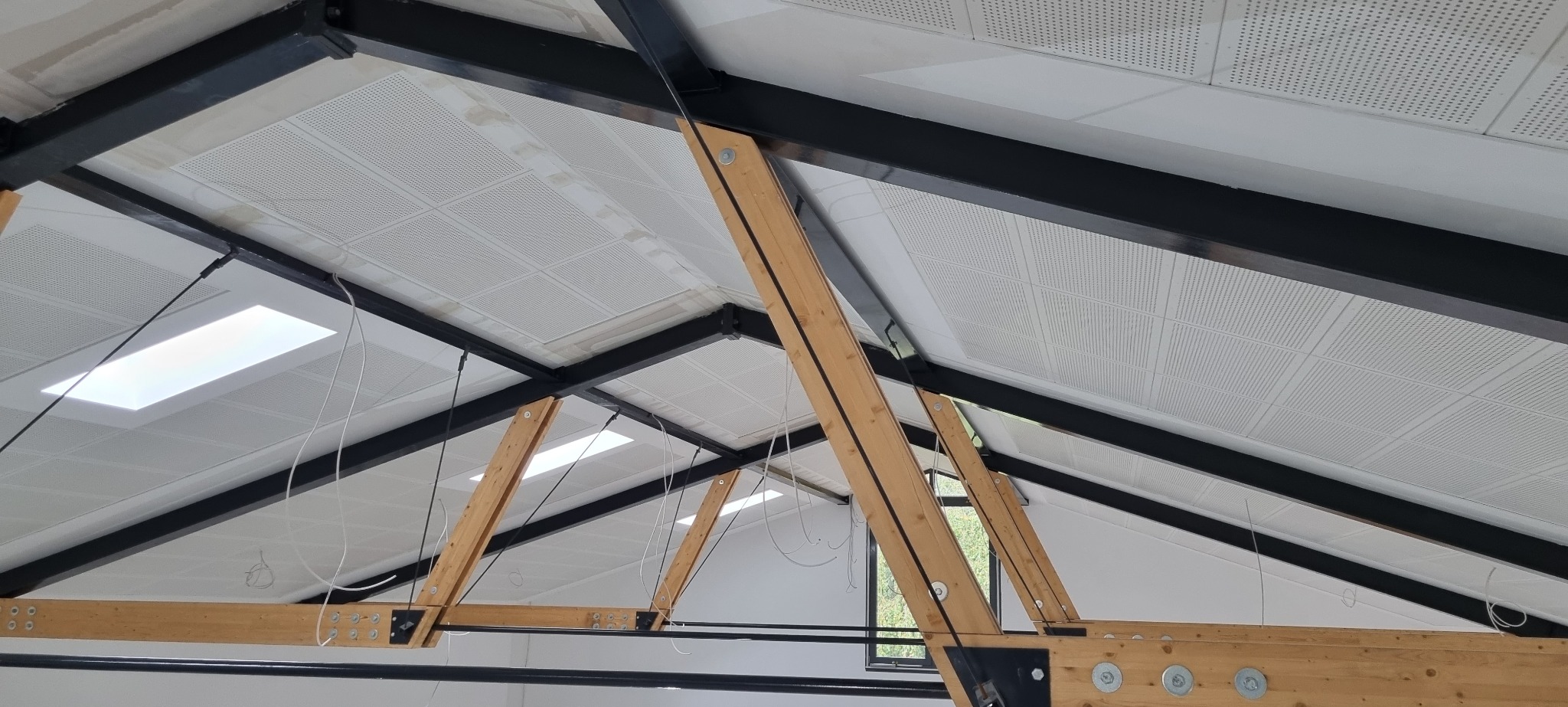 Montaż sufitu podwieszanego z perforowanych paneli na metalowej konstrukcji z drewnianymi wzmocnieniami, widoczne okna dachowe i przewody instalacyjne.