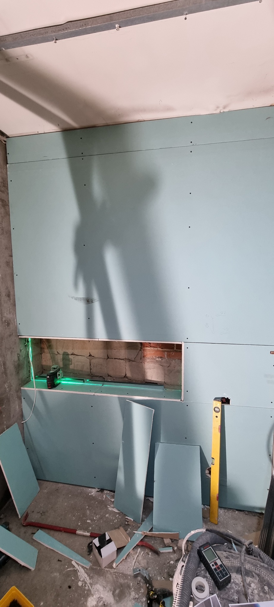 Montaż płyt gipsowo-kartonowych na ścianie z wnęką, poziomica laserowa wyznacza linię cięcia, na podłodze leżą fragmenty płyt i narzędzia.