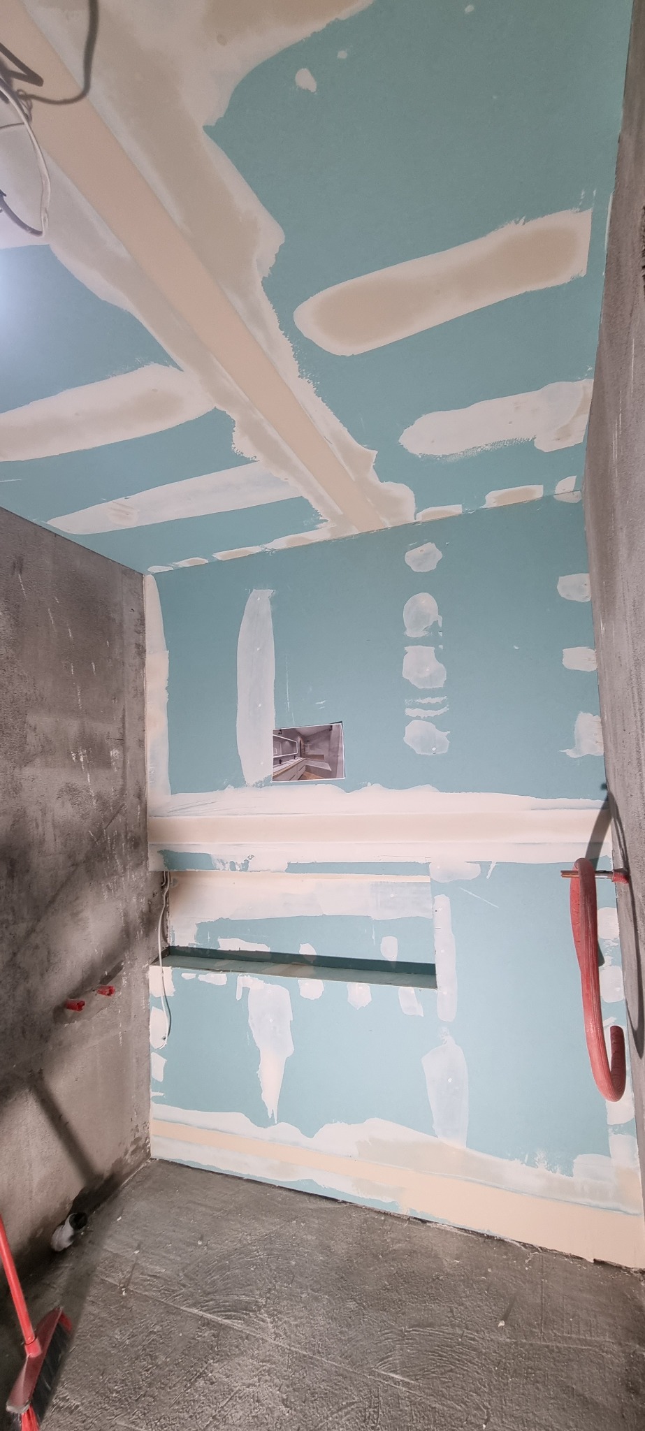 Niedokończona łazienka w trakcie remontu: ściany i sufit z niebieskich płyt gipsowo-kartonowych z widocznymi łączeniami zaszpachlowanymi na biało, surowa betonowa podłoga, widoczne instalacje wodne...