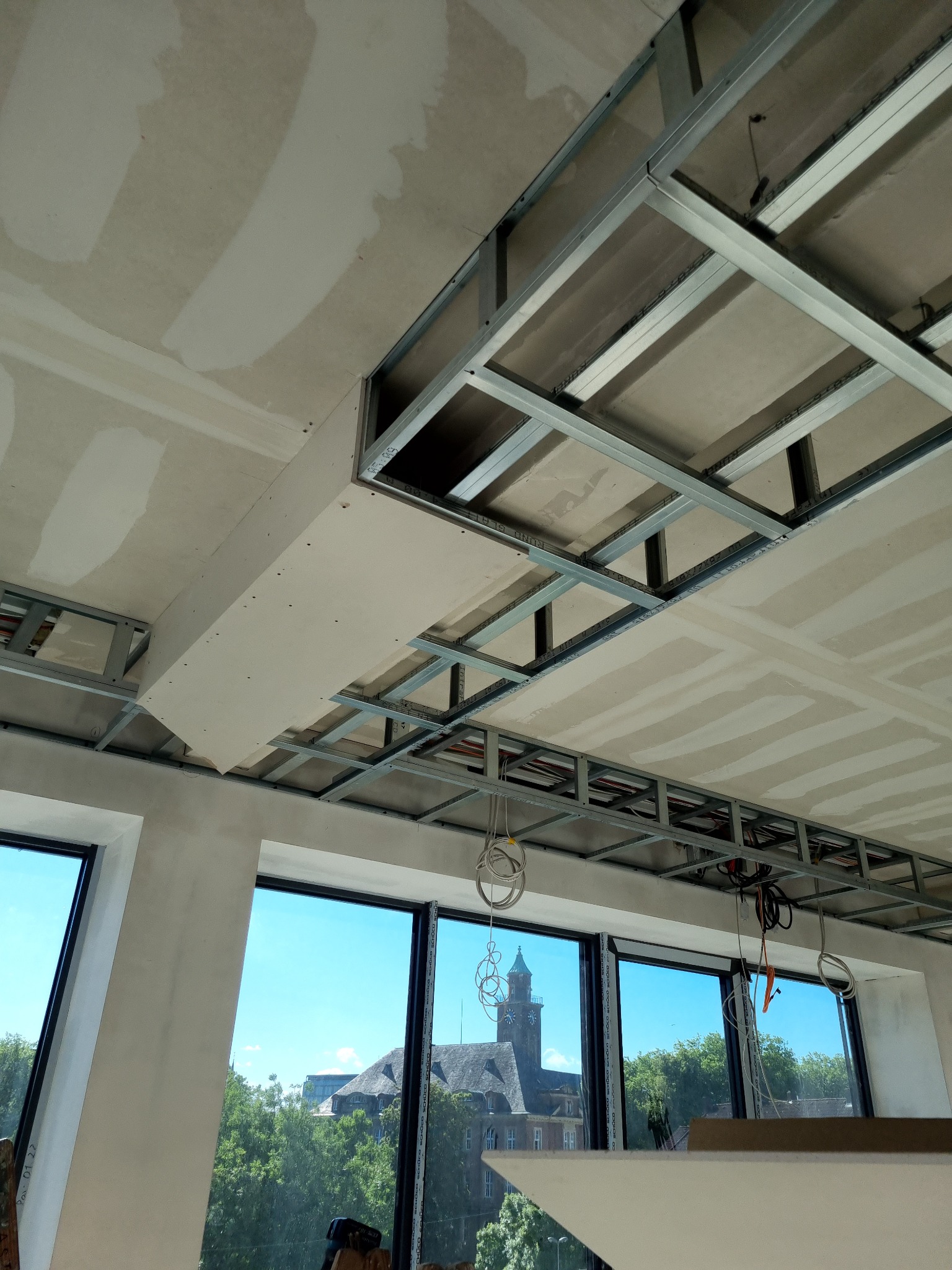 Montaż sufitu podwieszanego z metalowym stelażem i częściowo zamontowaną płytą, widok z dołu na okna z widokiem na budynek z wieżą zegarową.