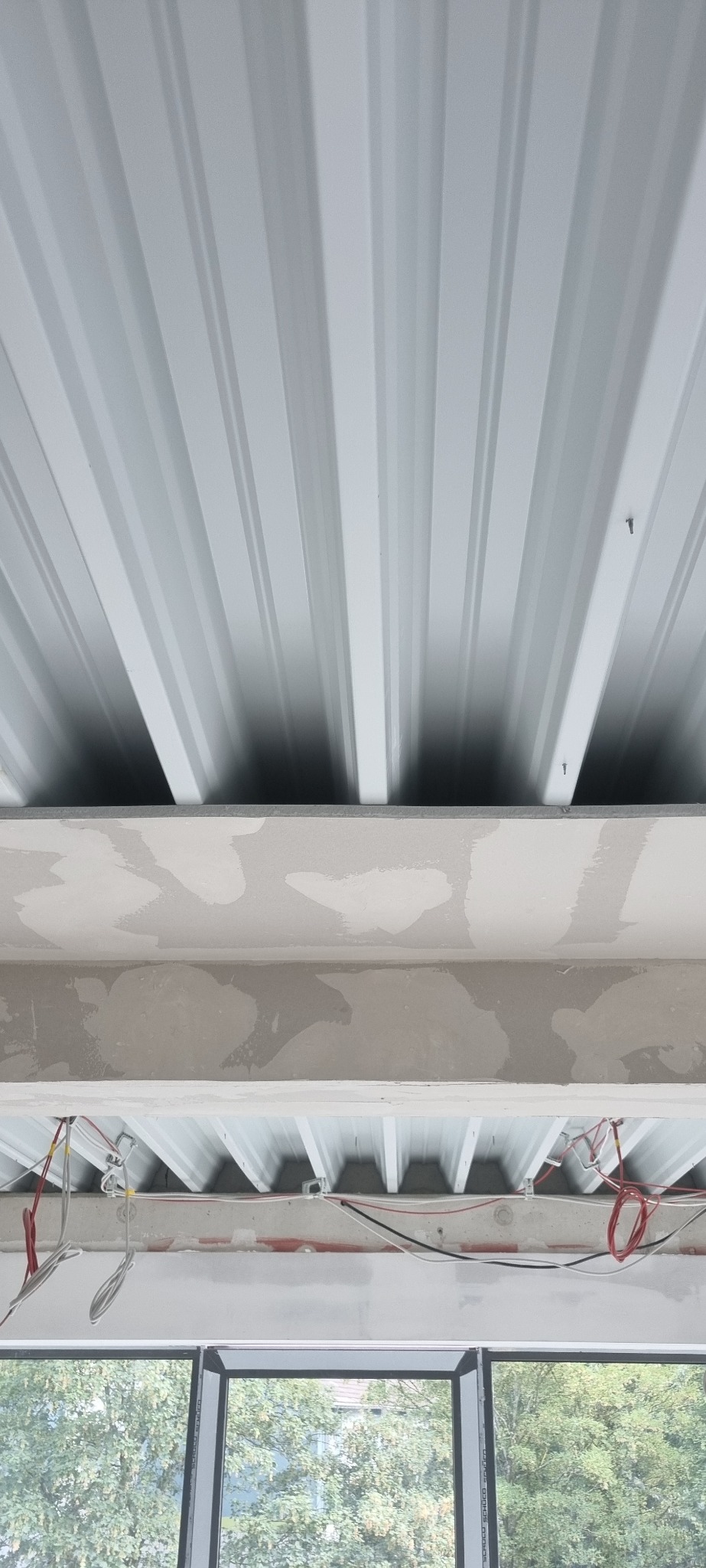 Sufit z blachy trapezowej, widoczne mocowania, poniżej sufit z niedokończonymi gładziami, wystające przewody elektryczne i okno z widokiem na drzewa.