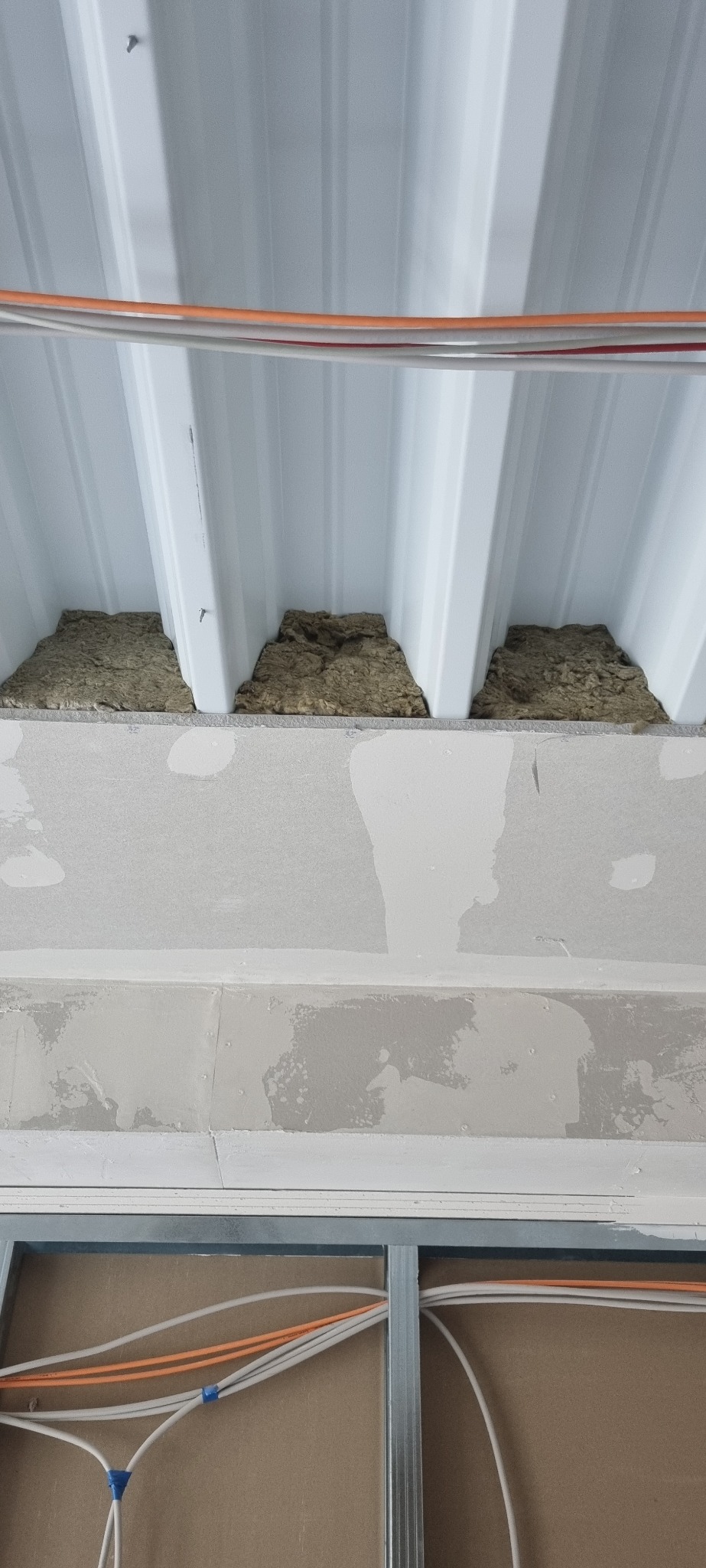 Wnętrze w trakcie prac wykończeniowych: izolacja wełną mineralną pod metalowym dachem, płyty gipsowo-kartonowe na ścianie, widoczne przewody elektryczne w metalowych profilach.