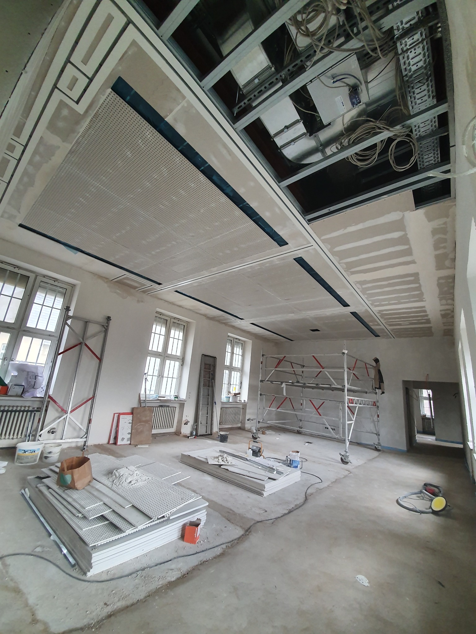 Wnętrze w trakcie montażu sufitu podwieszanego z płyt perforowanych. Widoczne rusztowania, płyty i instalacje pod sufitem. Okna z kratownicą w tle.