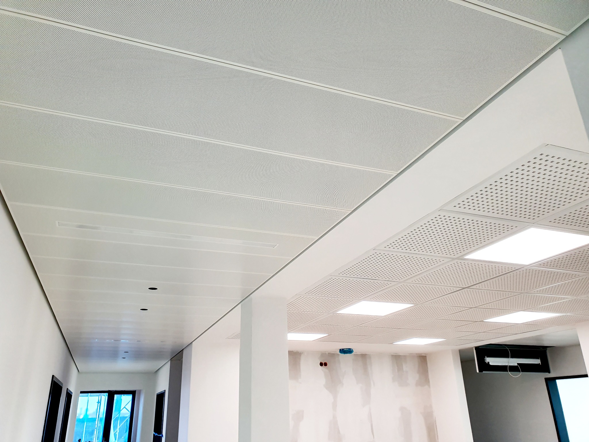 Nowoczesny sufit podwieszany z paneli perforowanych w jasnym wnętrzu biurowym. Widoczne elementy oświetleniowe i fragmenty ścian w trakcie wykończenia.