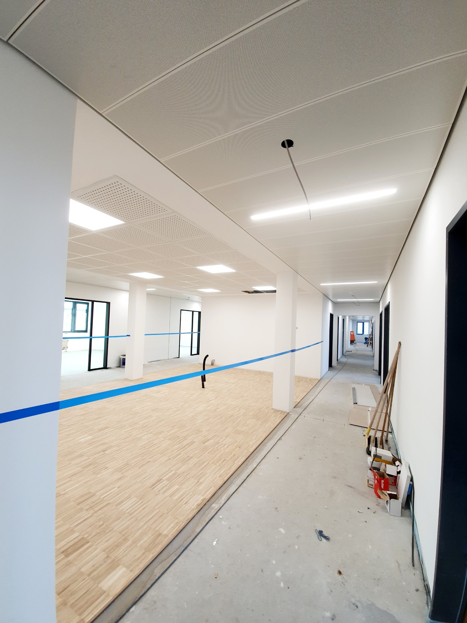 Wnętrze w trakcie wykończenia: drewniana podłoga częściowo ułożona, sufit podwieszany z oświetleniem. W tle widoczne drzwi i narzędzia, niebieska taśma wyznacza obszar robót.