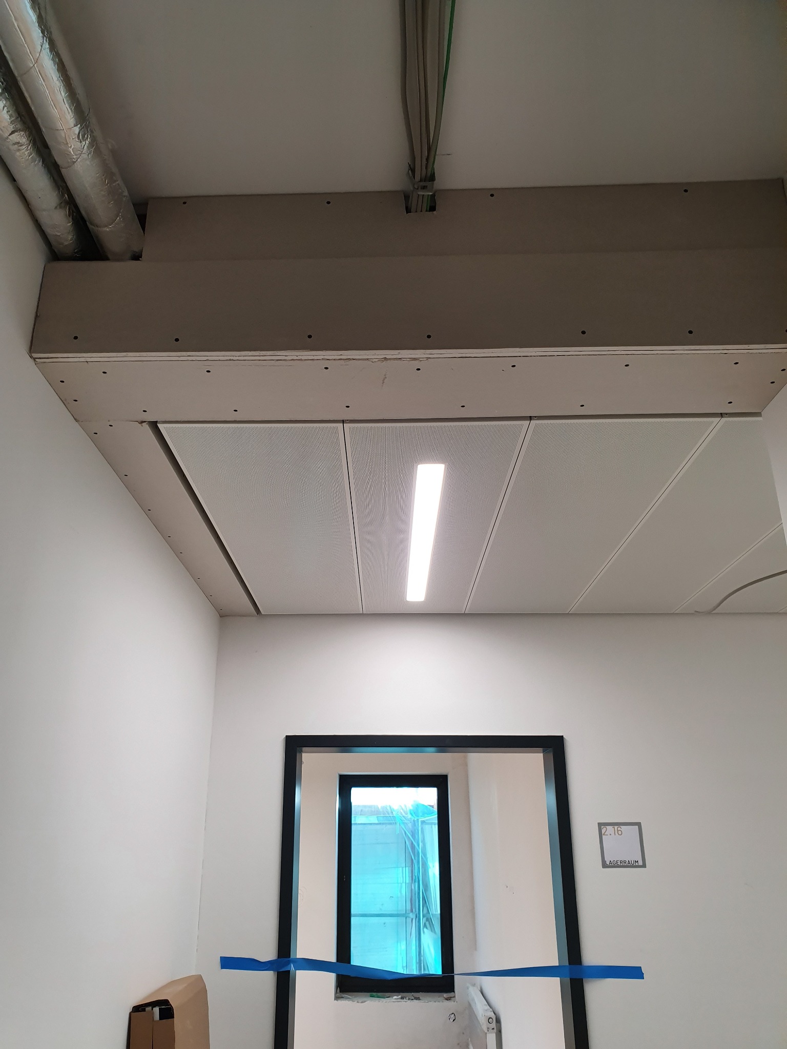 Wykończony sufit podwieszany z oświetleniem w Chocianowicach. Widoczne instalacje i fragment ściany z oknem. Minimalistyczny design, jasne wnętrze.