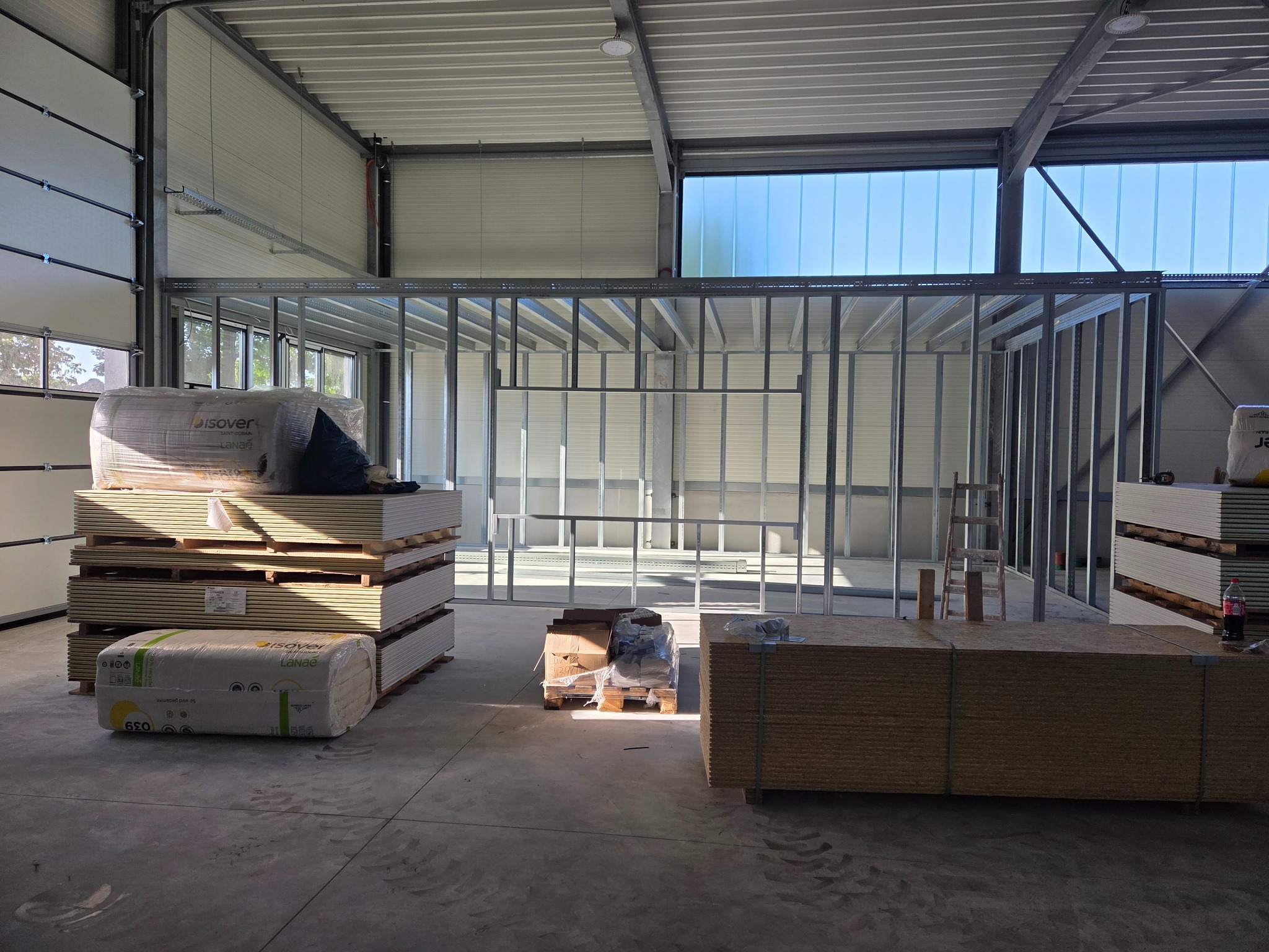 Wnętrze hali w trakcie prac: metalowa konstrukcja ścian działowych, palety z płytami i izolacją Isover, surowy beton na podłodze, okna w górnej części ściany.