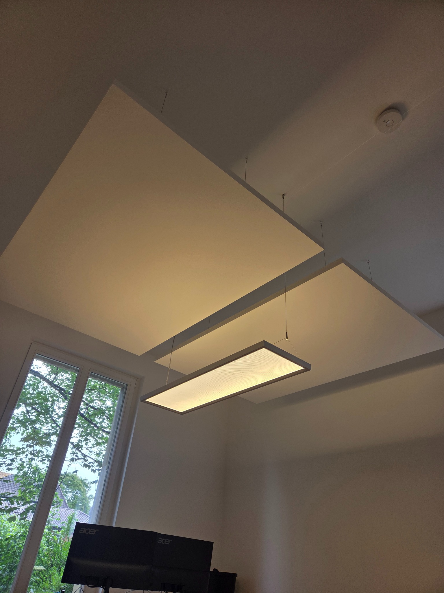 Nowoczesny sufit podwieszany z płyt w kształcie kwadratów, podświetlony wiszącą lampą. Widoczne okno z zielenią za szybą i czujnik dymu na suficie.