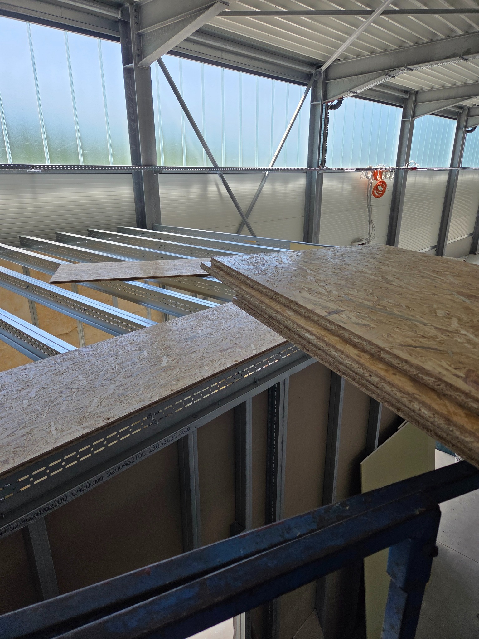 Konstrukcja poddasza z metalowych profili i płyt OSB, widok z góry. Częściowo ułożone płyty, surowe wnętrze budynku z oknami w tle. Etap prac wykończeniowych.