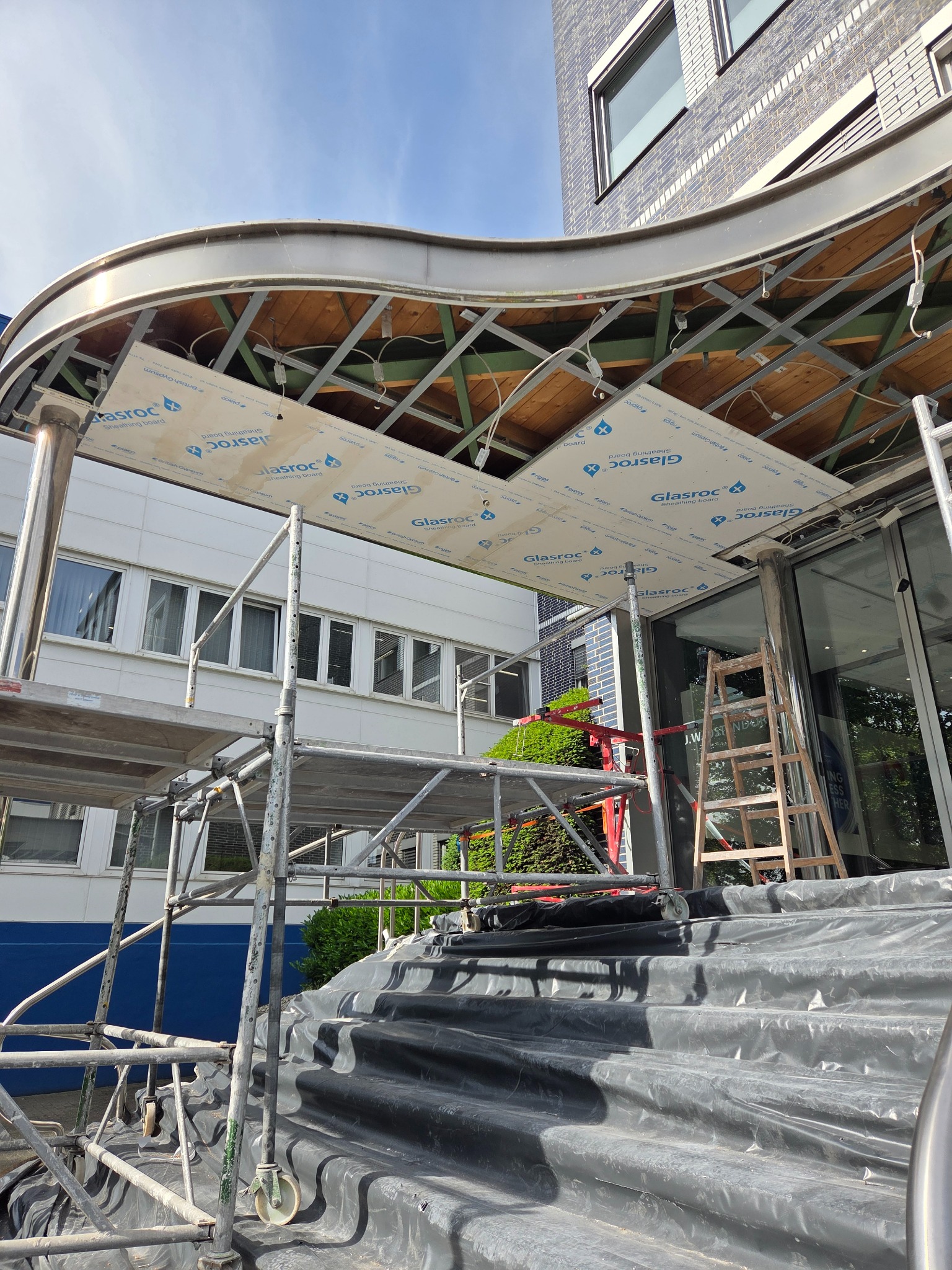 Montaż podbitki z płyt Glasroc pod zadaszeniem wejściowym budynku. Widoczne rusztowania i zabezpieczenia. Prace w toku, częściowo odsłonięta konstrukcja.