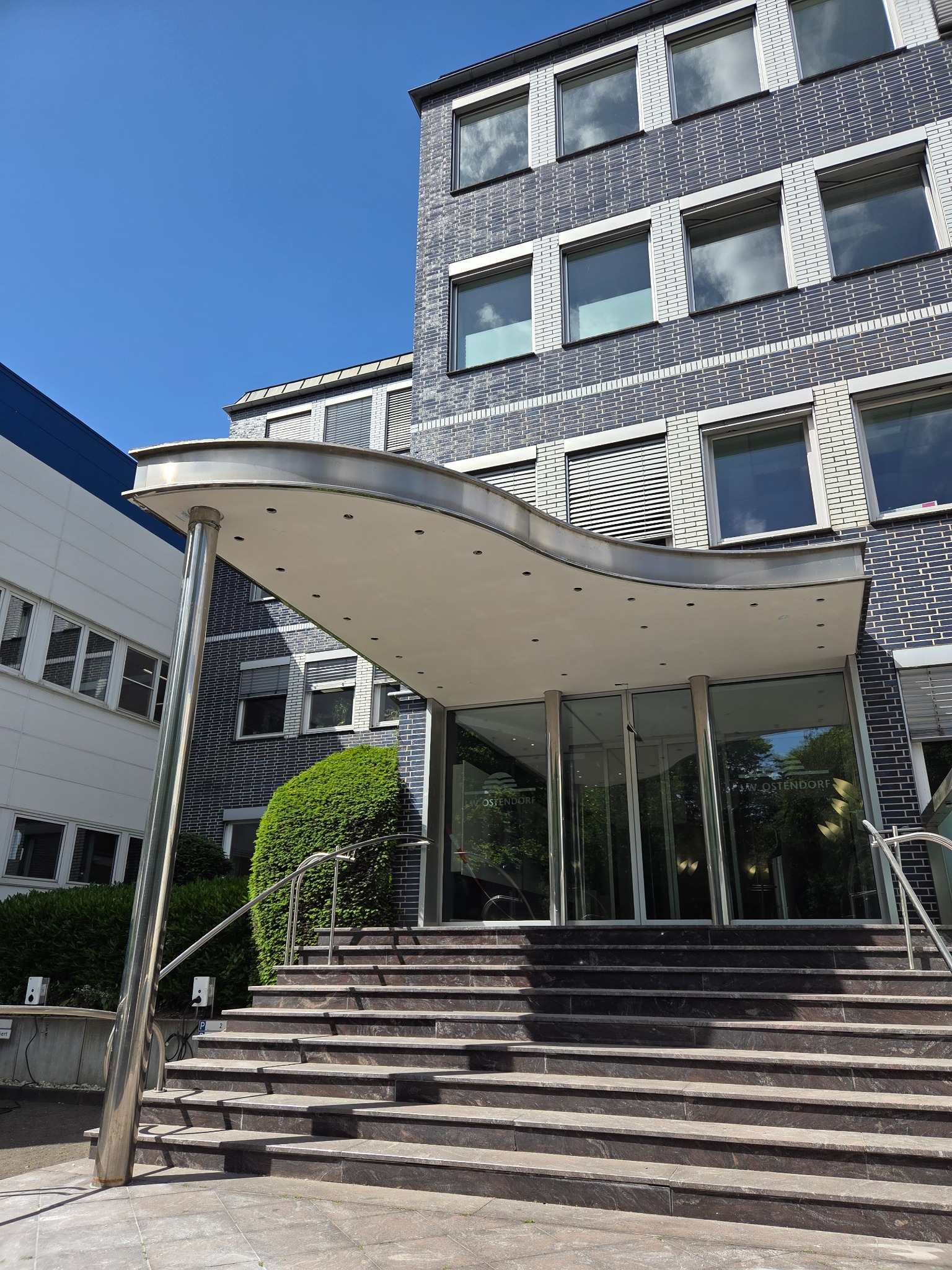 Elewacja budynku z wejściem, schodami i metalowym zadaszeniem. Budynek z szarej cegły z białymi obramowaniami okien. Błękitne niebo w tle.