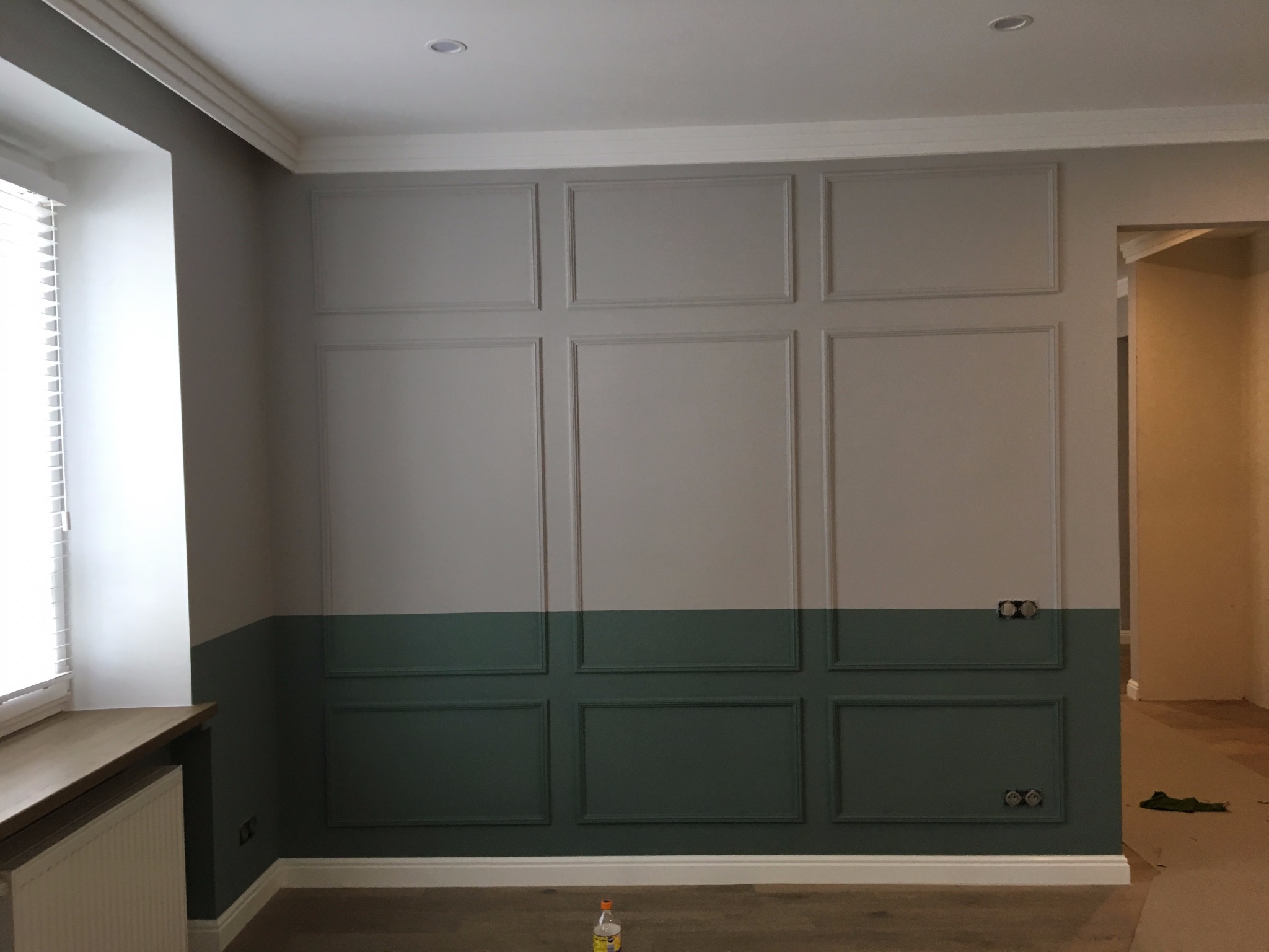 Ściana z dekoracyjnymi panelami w dwóch odcieniach szarości i zieleni, z widocznymi gniazdkami elektrycznymi i fragmentem okna z żaluzjami.