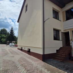 DAW-BUD - Dwukondygnacyjny dom z beżową elewacją i brązowymi akcentami, widoczny z boku. Na podjeździe leżą rusztowania. Słoneczny dzień, błękitne niebo z chmurami.