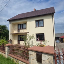 DAW-BUD - Dwupiętrowy dom z beżową elewacją, brązowym dachem i balkonami. Ozdobne ogrodzenie z kamiennymi słupkami i metalowymi przęsłami. W oknie widoczna osoba.