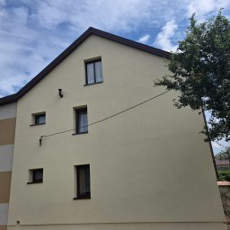 DAW-BUD - Elewacja domu po malowaniu w odcieniach beżu, z brązowymi ramami okiennymi i dachem. Widoczne okna o różnych rozmiarach i fragment drzewa po prawej stronie.