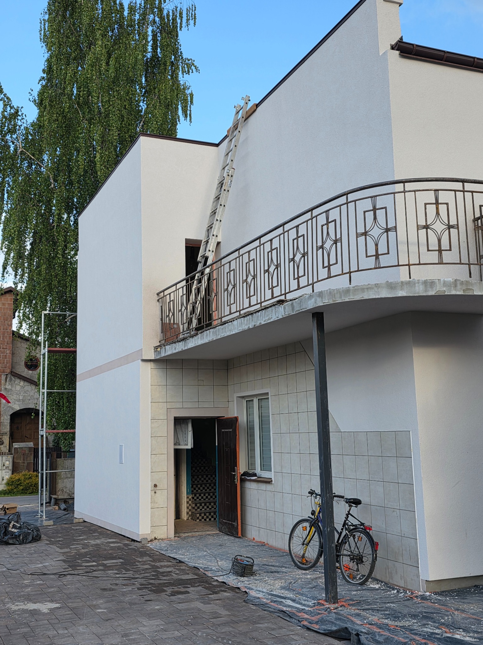 Dwupiętrowy dom z balkonem po remoncie elewacji, z drabiną opartą o balkon. Rower oparty o ścianę. Widoczne rusztowania i zabezpieczenia na podłożu.