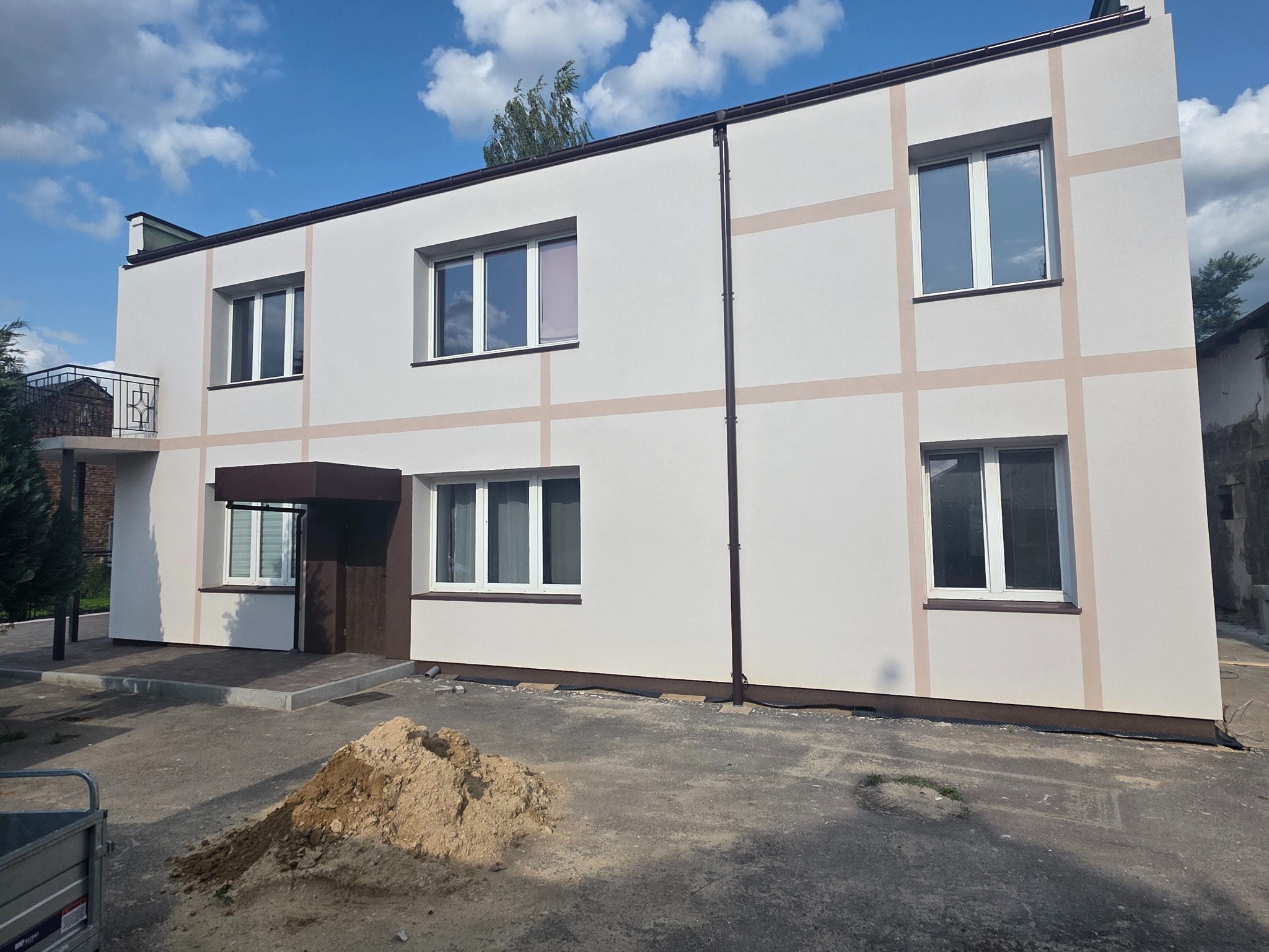 Dwukondygnacyjny budynek po ociepleniu, z beżowymi pasami na elewacji, brązowymi obramowaniami okien i dachem. Na pierwszym planie hałda piasku.