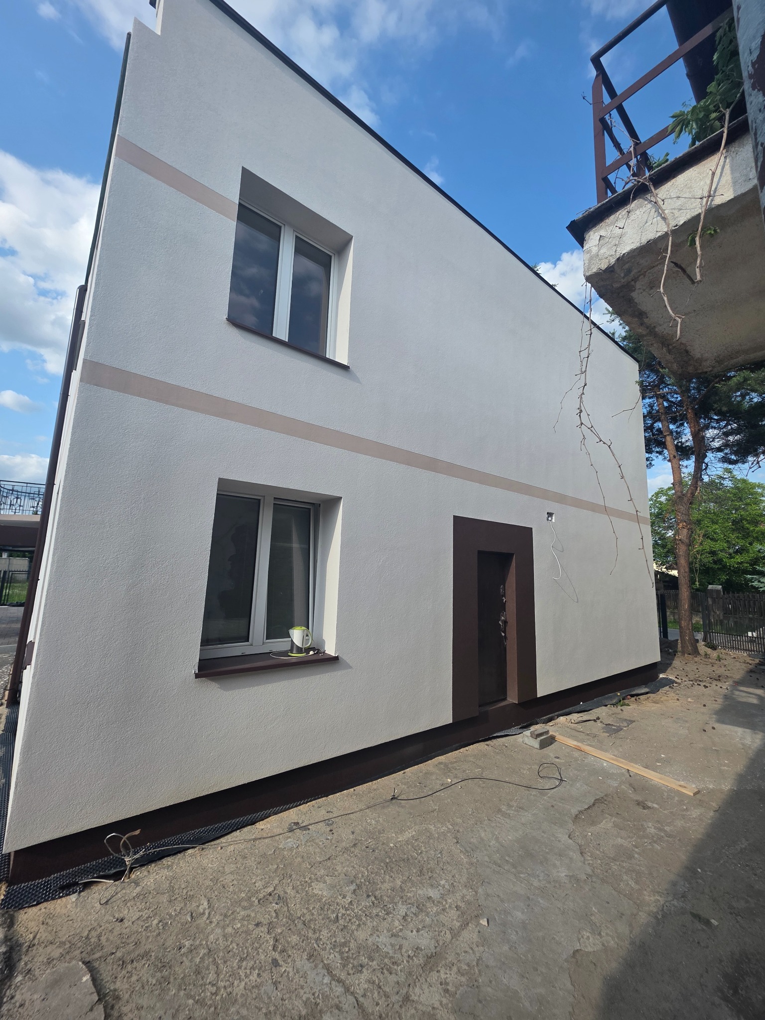 Biała elewacja domu z brązowymi akcentami: okna, drzwi i pasy dekoracyjne. Widoczne fragmenty balkonu i drzew. Ujęcie z dołu, podkreślające wysokość budynku.