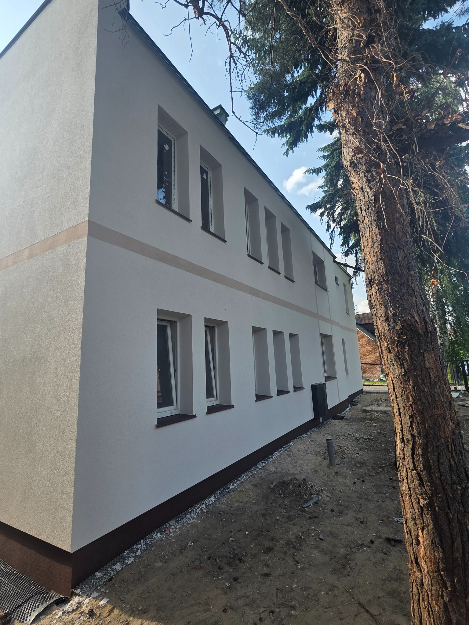 Biała elewacja budynku z rzędem okien, wykończona poziomym pasem w odcieniu beżu. Obok wysokie drzewo iglaste, błękitne niebo w tle. Ujęcie z dołu.