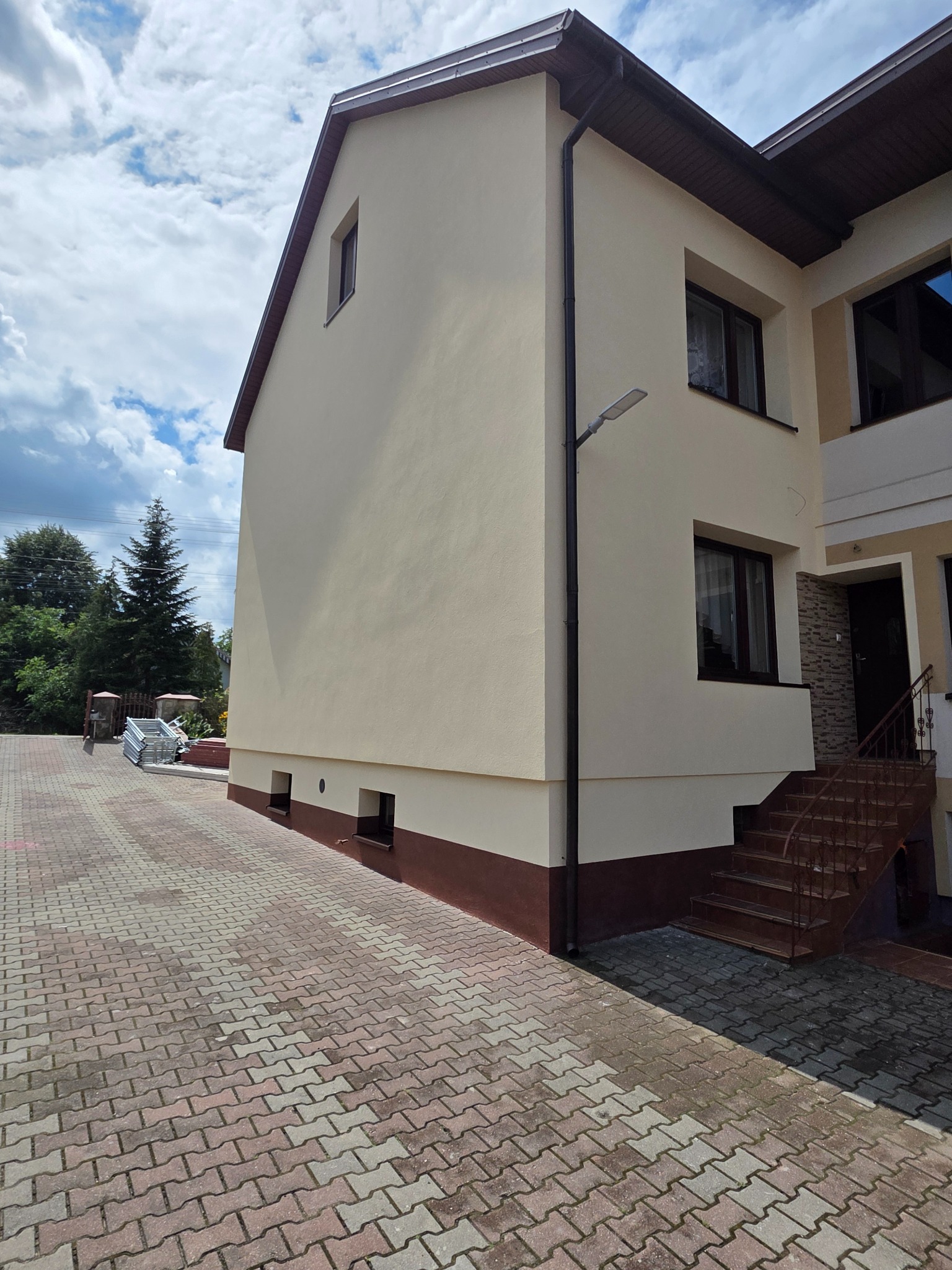 Dwukondygnacyjny dom z beżową elewacją i brązowymi akcentami, widoczny z boku. Na podjeździe leżą rusztowania. Słoneczny dzień, błękitne niebo z chmurami.