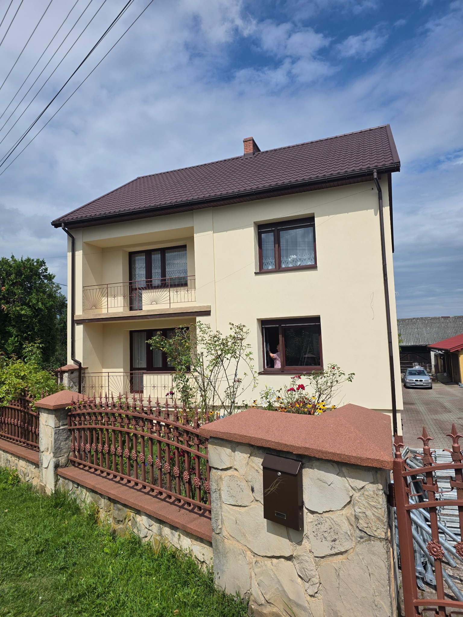 Dwupiętrowy dom z beżową elewacją, brązowym dachem i balkonami. Ozdobne ogrodzenie z kamiennymi słupkami i metalowymi przęsłami. W oknie widoczna osoba.